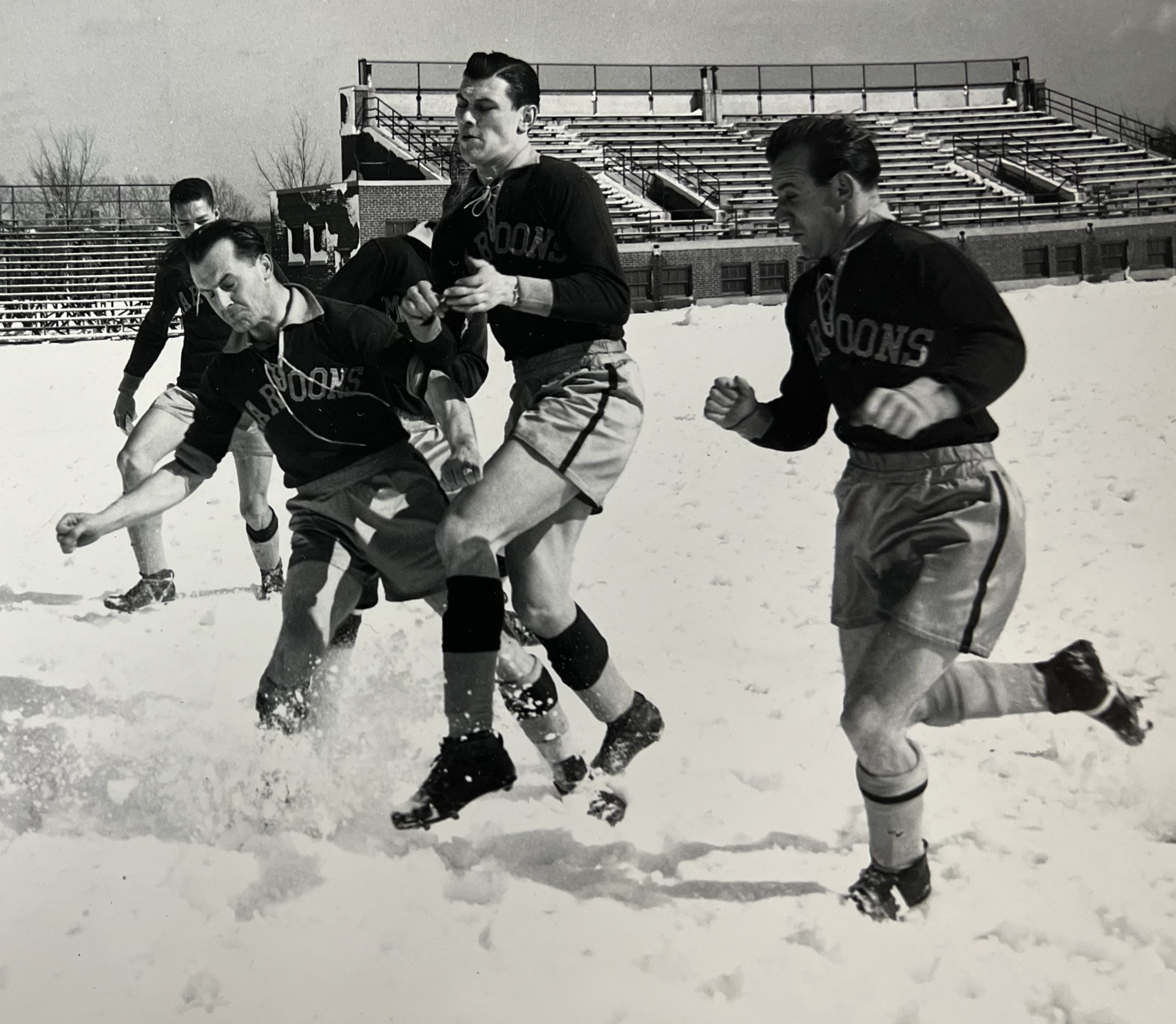 5 soccer players from Chicago Maroons playing soccer in snow with stands in background